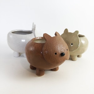 May include: Three ceramic animal planters in a row. The front planter is brown with a black nose and small ears. The middle planter is a light brown color. The back planter is white with antlers.