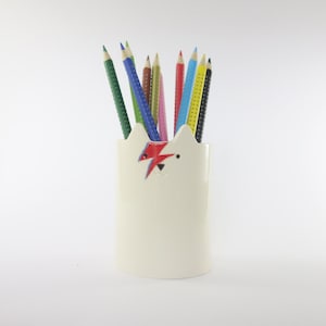 May include: White ceramic pencil holder shaped like a cat's head with a red and blue lightning bolt design on the forehead. The holder is filled with colored pencils.