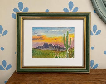 Può includere: Un dipinto ad acquerello incorniciato che raffigura un paesaggio desertico al tramonto. L'opera presenta un alto cactus saguaro verde, montagne viola scuro e un cielo vibrante con tonalità gialle e arancioni, racchiuso in una cornice verde e oro.