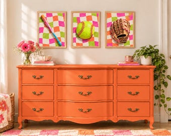 Può includere: Una cassettiera in legno color arancione brillante con nove cassetti e pomelli in metallo decorati, posizionata sotto tre stampe da parete incorniciate raffiguranti una mazza da softball, una pallina da softball gialla e un guanto da baseball in pelle marrone su sfondo a scacchi rosa e verde.