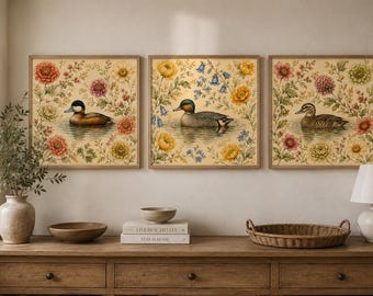 Può includere: Un set di tre stampe d'arte da parete quadrate incorniciate, ognuna raffigurante un'anatra che nuota nell'acqua circondata da colorate illustrazioni floreali nei toni del rosso, giallo e verde. Le stampe sono in cornici di legno chiaro.