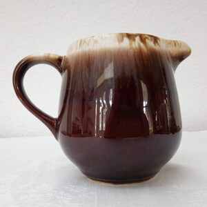 May include: A brown ceramic creamer with a white glaze. The creamer has a rounded body and a handle.