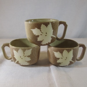 May include: Three brown ceramic mugs with white leaf designs. The mugs are stacked on top of each other.