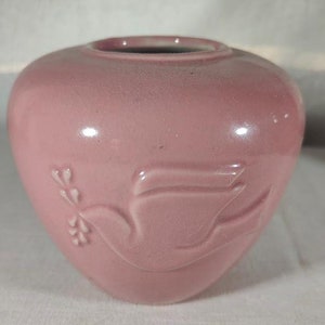May include: A pink ceramic vase featuring a dove design. The vase is heart-shaped and has a smooth, glossy finish.