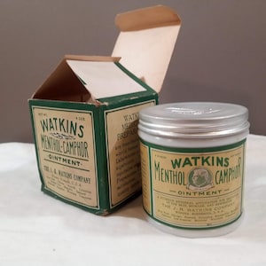 Puede incluir: Una caja verde y blanca y un frasco blanco con tapa plateada. Tanto la caja como el frasco tienen el texto "Watkins Menthol-Camphor Ointment". La caja está abierta y el frasco está sobre una superficie blanca.