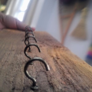 May include: A close-up of a wooden board with a row of metal hooks. The hooks are all in a row and are evenly spaced. The wood is a dark brown color and has a rough texture.
