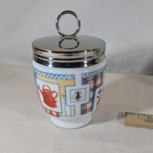Può includere: Un portauovo in ceramica bianca con un coperchio argentato e un design patchwork colorato con un'ape, un annaffiatoio e motivi a quadri.