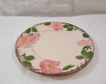 May include: A white ceramic plate with a pink rose pattern. The plate has a brown rim and green leaves around the roses.