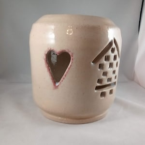 Può includere: Un portacandele in ceramica beige con un design a forma di cuore e casa ritagliati. I ritagli sono contornati da un colore marrone rossastro.
