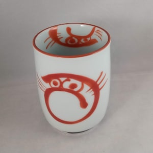 May include: White ceramic teacup with a red and orange stylized cat design. The cat design is repeated on the inside rim of the cup.