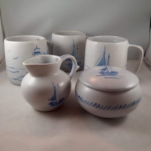 May include: A collection of ceramic tableware with a nautical theme. The set includes mugs, a creamer, and a lidded jar, all in a speckled white finish. Each piece is decorated with blue sailboat and lighthouse designs, evoking a coastal aesthetic.