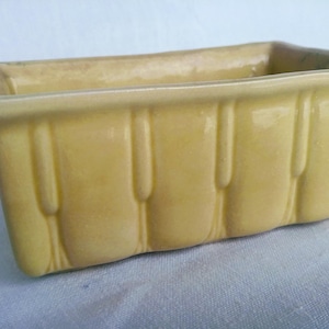 Puede incluir: Un macetero de cerámica amarillo con forma rectangular y un diseño texturizado. El macetero tiene un acabado liso y brillante.