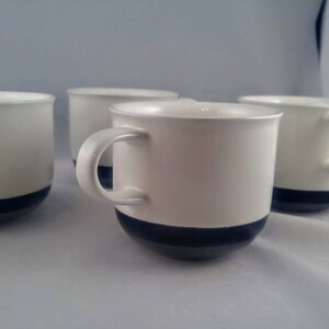 Op de afbeelding: Vier witte keramische koffiemokken met een zwarte band rond de bovenkant. De mokken hebben een eenvoudig ontwerp en zijn perfect voor dagelijks gebruik.