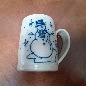 Könnte beinhalten: Eine weiße Keramiktasse mit einem blauen Schneemann-Design. Der Schneemann trägt einen schwarzen Hut und einen Schal. Um den Schneemann herum sind Schneeflocken.