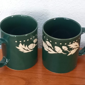 May include: Two green ceramic mugs with white acorn and leaf designs. The mugs have a white dotted line around the top.