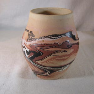 Puede incluir: Un jarrón de cerámica hecho a mano con una base ancha y un borde ligeramente ensanchado. El jarrón presenta un patrón arremolinado de colores marrón, negro, blanco y beige. La forma general es redondeada y tiene una estética natural y terrosa.