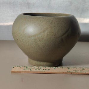 May include: A speckled, olive-green ceramic bowl with a rounded body and a small, circular base. The bowl has a smooth, matte finish and a subtle, leaf-like design. A wooden ruler is in the foreground.