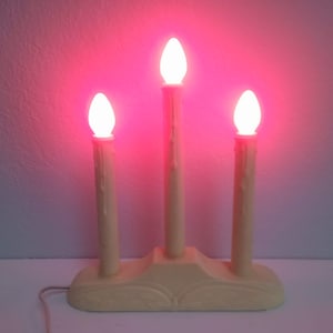 May include: A set of three white candle-shaped light bulbs with a pink glow, mounted on a beige wooden base. The base has a decorative design and a cord for plugging in.