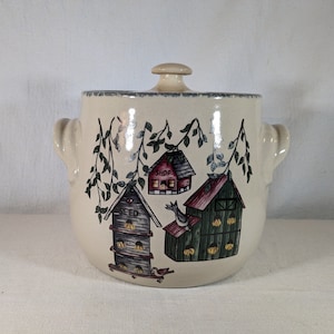 May include: A ceramic crock with a lid, featuring a whimsical design of birdhouses with the words "Shop", "Seed", and "Feed" on them. The crock is decorated with green vines and birds.