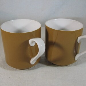 May include: Two brown ceramic mugs with white interiors and white handles. The mugs are sitting on a white surface.