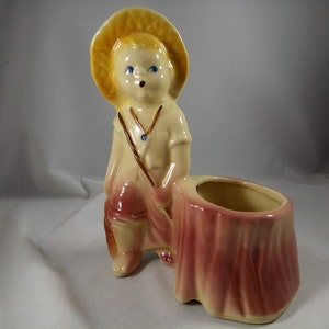 Puede incluir: Un macetero de cerámica con forma de niño que lleva un sombrero de paja y sostiene una caña de pescar. El macetero está sentado sobre un tocón de árbol de cerámica rosa y amarillo.