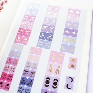 Könnte beinhalten: Blatt mit Planer-Aufklebern mit monatlichen Etiketten, mit goldenen Folienakzenten und Himmelsmotiven. Die Aufkleber sind in Rosa-, Lila- und Blautönen gehalten. Die Etiketten enthalten Monate wie Mai, Juni und November.