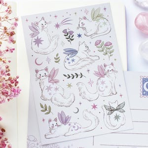 May include: A sheet of stickers and postcards featuring whimsical illustrations of white cats with fairy wings, stars, and moons. The stickers and postcards have a pastel color palette with pink, purple, and green accents. A sprig of pink flowers is in the background.