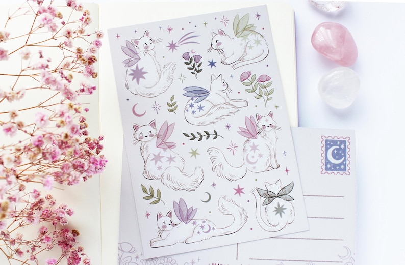 May include: A sheet of stickers and postcards featuring whimsical illustrations of white cats with fairy wings, stars, and moons. The stickers and postcards have a pastel color palette with pink, purple, and green accents. A sprig of pink flowers is in the background.