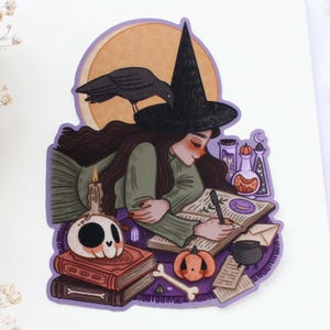 May include: A sticker illustration of a person with long dark hair wearing a black witch hat, writing in an open book. A black crow sits on the hat. Surrounding elements include a skull, stacked books, a candle, potions, and a pumpkin.