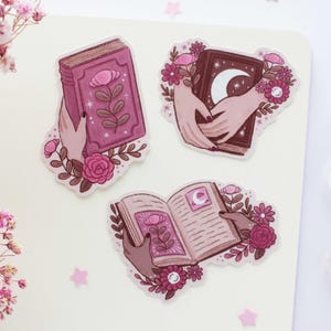 Könnte beinhalten: Drei rosafarbene und braune illustrierte Aufkleber mit einem geschlossenen Buch mit einer Blume, einem offenen Buch mit einer Mondsichel und einer Hand, die ein Buch mit einer Mondsichel hält.