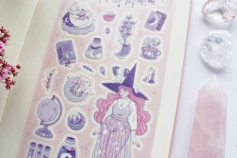 Puede incluir: Una hoja de pegatinas con una bruja de pelo rosa y un sombrero morado, un libro con el texto "Moon Magic Spells", un gato en una nube, una botella de poci&oacute;n y otros elementos m&aacute;gicos. Las pegatinas est&aacute;n sobre un fondo rosa.