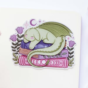 Puede incluir: Una pegatina con un dragón verde durmiente sobre una pila de libros. Los libros son morados y rosas, con detalles florales y celestiales. El dragón tiene los ojos cerrados y una larga cola. La pegatina está rodeada de flores moradas y una luna creciente.