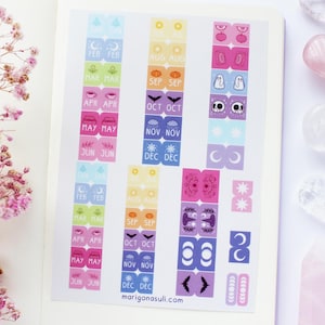 Peut inclure: Une feuille d'autocollants de planificateur colorés avec divers motifs, dont des lunes, des yeux, des chauves-souris et des fantômes. Les autocollants sont disposés en colonnes et présentent des mois comme mai, juin et octobre. Le site Web moriganasuli.com est visible.