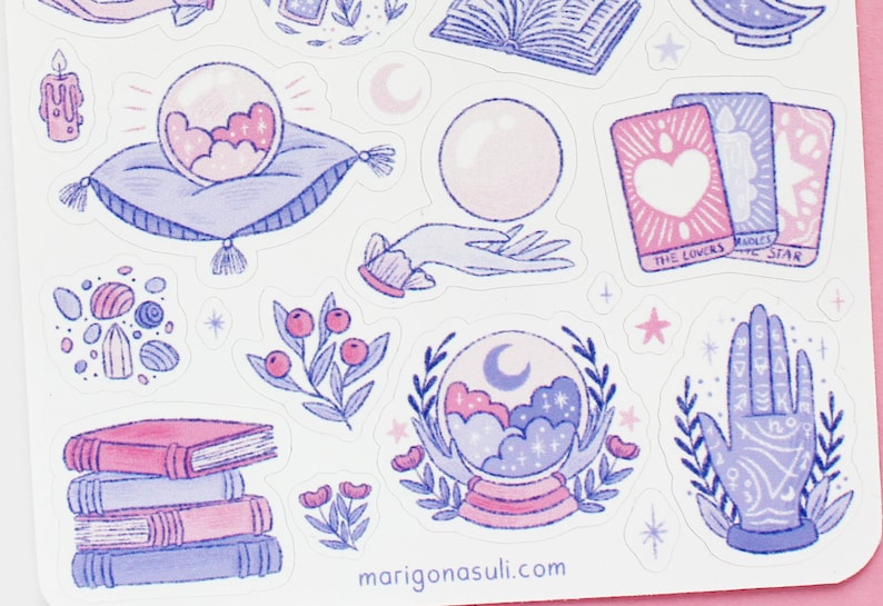 May include: A sheet of stickers featuring mystical and celestial illustrations in shades of pink, purple, and blue. Designs include a crystal ball, tarot cards, a hand with symbols, books, and floral elements. The website address marigonasuli.com is visible.