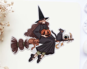 May include: A die-cut sticker featuring a character in a black pointed hat and long dark robe riding a broomstick. The character holds an orange jack-o'-lantern, accompanied by a brown cat, a black cat, a small skull, and a lit candle.