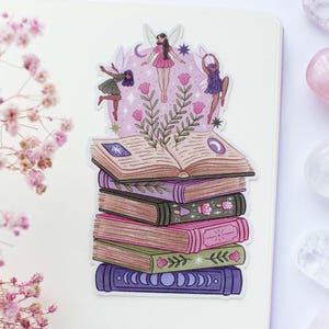 May include: A sticker depicting a stack of books with a fairy and floral design. The books are in shades of pink, green, and purple. The top book is open, and the sticker has a whimsical, fantasy theme.