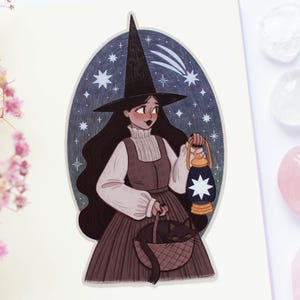May include: An illustration of a witch with long black hair, wearing a tall black hat and holding a lantern with a star design. She carries a basket with a black cat. The background is a dark blue oval with stars.