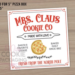 May include: A square white label with red and black text and illustrations. The label reads "MRS. CLAUS COOKIE CO" with a rolling pin graphic and "MADE WITH LOVE." It also says "SANTA'S FAVORITE COOKIE" and "BAKED DAILY SINCE 1882." The label is sized for a 5" x 5" pizza box.
