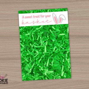 May include: A rectangular card with the text "A sweet treat for your basket" and bunny ears graphic, set against a background of bright green shredded paper. The card is white with pink text and a gray bunny ears graphic.