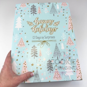 May include: A light blue holiday-themed box with the words "happy holidays" and "12 Days of Surprises" in gold script. The box is decorated with illustrations of Christmas trees, stars, and snowflakes in white, pink, and gray.