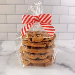 May include: A stack of chocolate chip cookies in a clear cellophane bag, tied with a red and white striped ribbon. The cookies are golden brown and appear freshly baked, with visible chocolate chips. The background is a white brick wall.