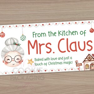 May include: A white rectangular label with a red and white scalloped border. The label features a cartoon illustration of Mrs. Claus with a red bow in her hair. The text "From the kitchen of Mrs. Claus" is in red and green lettering. The text "Baked with love and just a touch of Christmas magic!" is in black lettering.