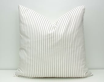 May include: A square cushion with a white base and vertical grey stripes. The cushion is positioned on a white surface against a white wall. The cushion has a soft, inviting appearance, suitable for home decor.