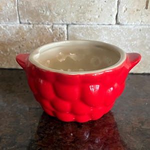 May include: A small, red ceramic bowl with a textured exterior and two small handles. The interior is a light beige color. The bowl is sitting on a dark surface.