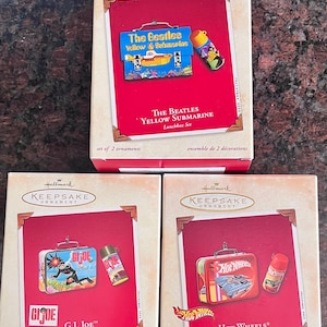 May include: Three Hallmark Keepsake Ornament boxes featuring lunchbox sets. The sets include The Beatles Yellow Submarine, G.I. Joe, and Hot Wheels designs. Each box contains two ornaments. The boxes have a red background with the ornament images.