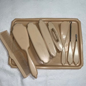 May include: A set of vintage, off-white, plastic hair care tools in a matching tray. The set includes a comb, a hairbrush, a nail file, a cuticle pusher, and two brushes.