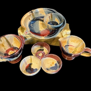 May include: A handmade ceramic serving set with a variety of bowls and a pitcher. The set features a color palette of yellow, red, blue, and black. The bowls have a unique, organic shape and are suitable for serving snacks or dips.
