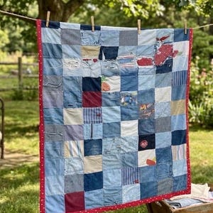 May include: A patchwork quilt made from repurposed denim squares, featuring various shades of blue and denim textures. The quilt is bordered with a red fabric and is hanging on a clothesline with wooden clothespins. The quilt is a handmade item.