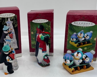 May include: Set of three Hallmark Keepsake Christmas ornaments featuring penguins. The first ornament shows two penguins decorating a Christmas tree. The second ornament shows two penguins wearing scarves and hats. The third ornament shows three penguins riding a sled.