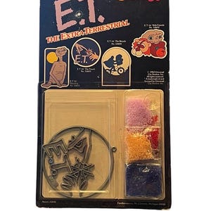May include: A vintage Makit & Bakit E.T. The Extra-Terrestrial stained glass suncatcher craft kit in original packaging. The set includes a circular metal frame, small bags of pink, red, yellow, and blue plastic crystals, and a suction cup.